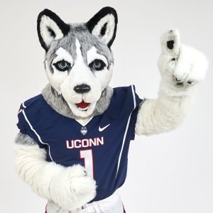 Studio portrait of Jonathan the Husky mascot wearing a UConn football jersey, posing with one finger raised.