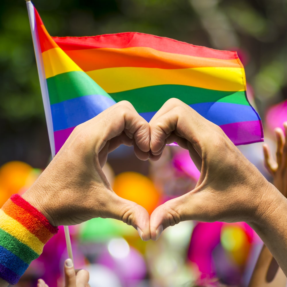 LGBTQIA love graphic: Hands forming a heart shape in front of a rainbow pride flag during a colorful outdoor event.