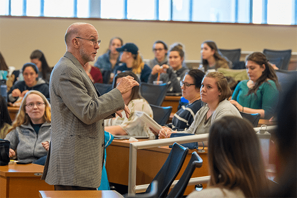 An older male professor stands in front of a lecture hall full of students who are seated and attentively listening to him.