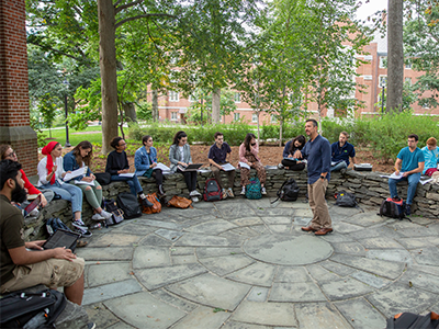 Group of students seated in a circle outdoors engaged in a discussion with a standing instructor.