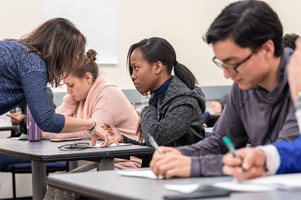 A teacher leans over a student’s desk offering guidance while other students write at their desks in a classroom setting.