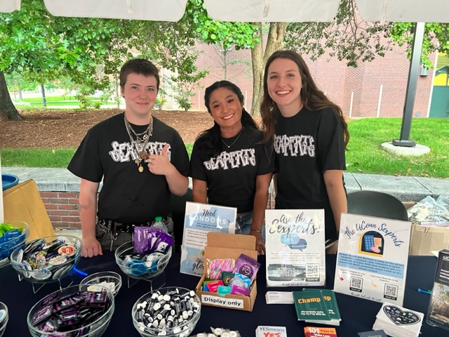 Three people stand behind a table outdoors with a black tablecloth displaying wellness products and informational signs.