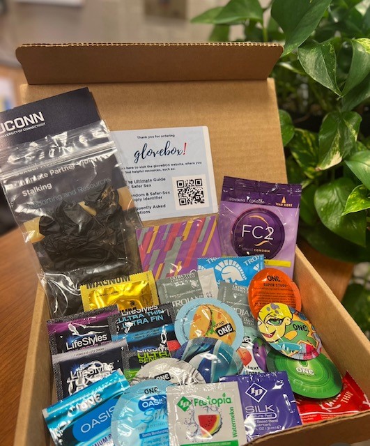 Box of assorted safer sex supplies including condoms and informational cards, displayed open on a table near a plant.