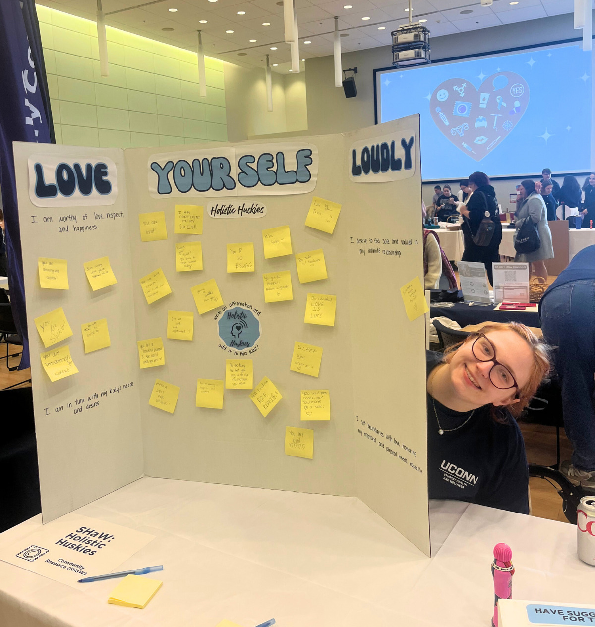 Holistic Huskies Event A student smiles next to a “Love Yourself Loudly” board covered in sticky notes with affirmations promoting self-love and wellness.