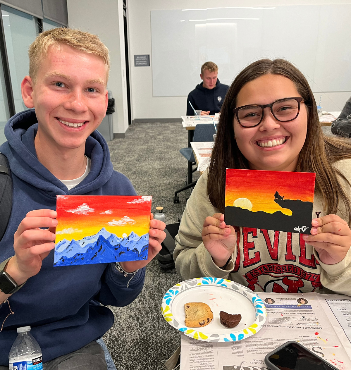 Holistic Huskies painting1 Two students proudly display their vibrant sunset paintings while enjoying snacks.