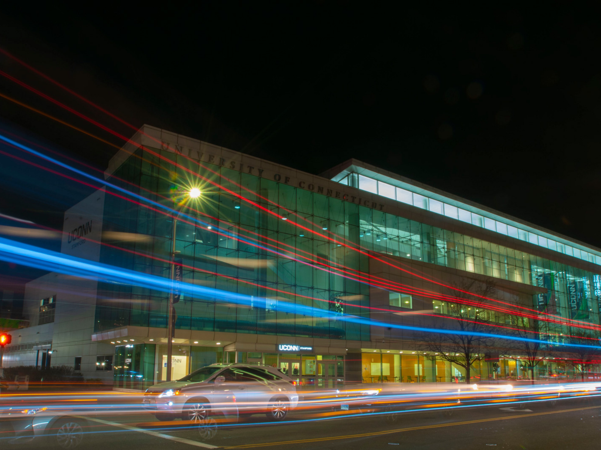 Nighttime exterior of UConn Stamford’s modern glass campus building with light trails from passing cars in the foreground.