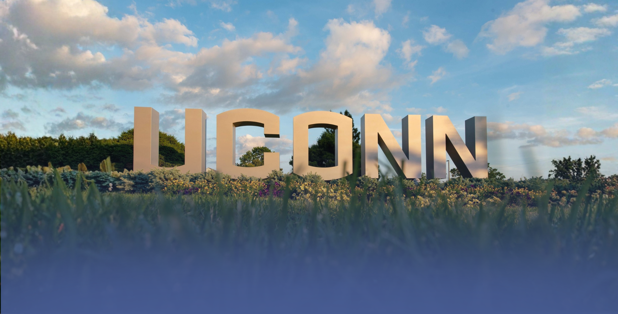 Large white “UCONN” letters stand outdoors in a landscaped area with flowers and greenery, set against a blue sky with scattered clouds.