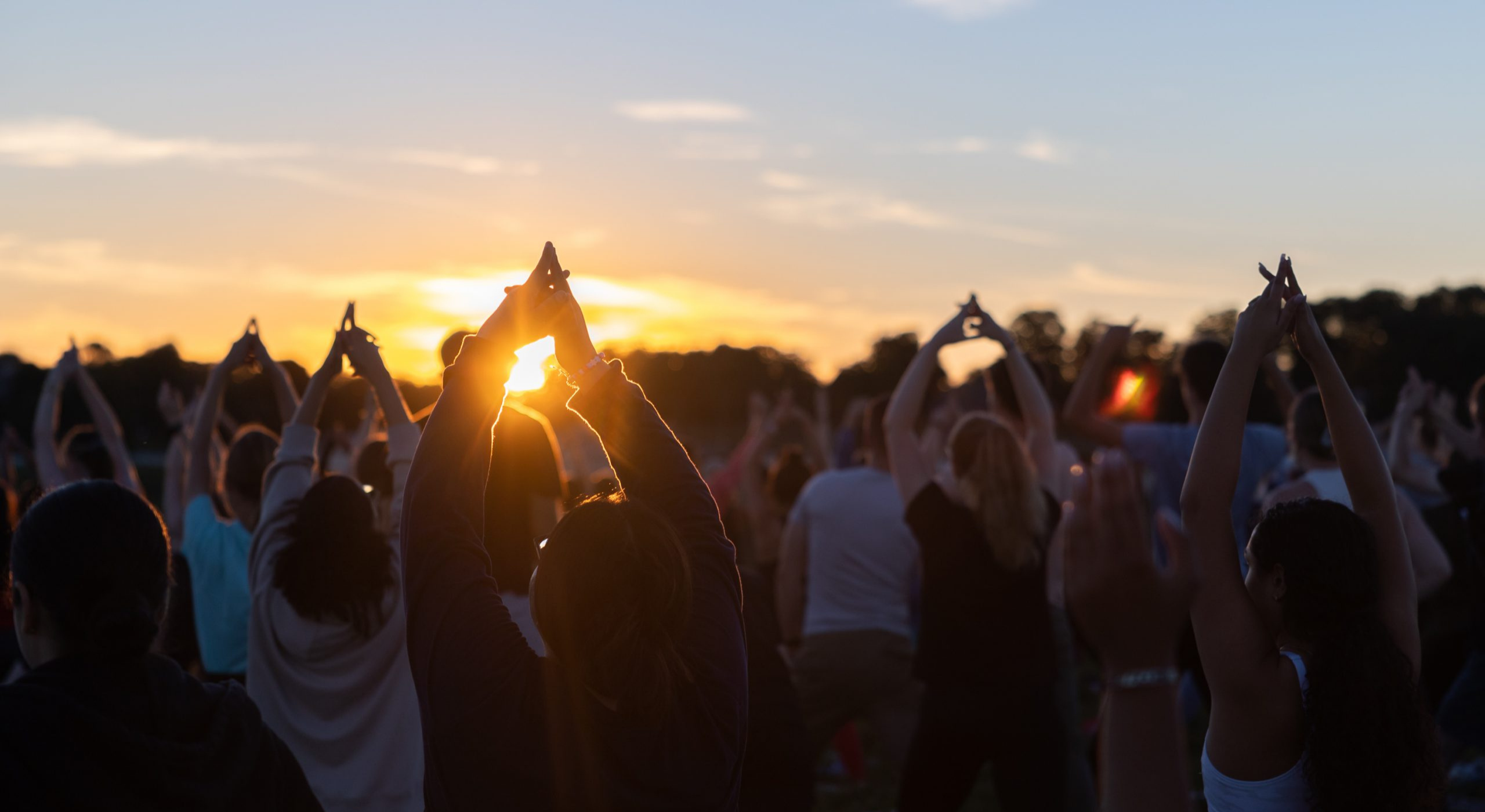 A group of people participate in an outdoor yoga session at sunset, with arms raised as the sun sets behind them