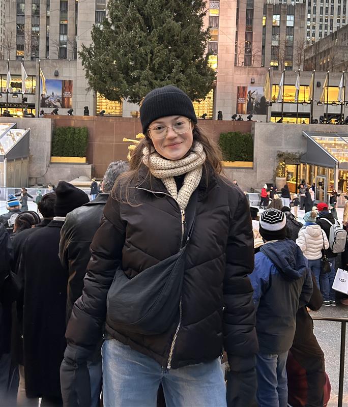 Student stands outdoors in a busy city plaza during winter, wearing a coat and scarf among a crowd.