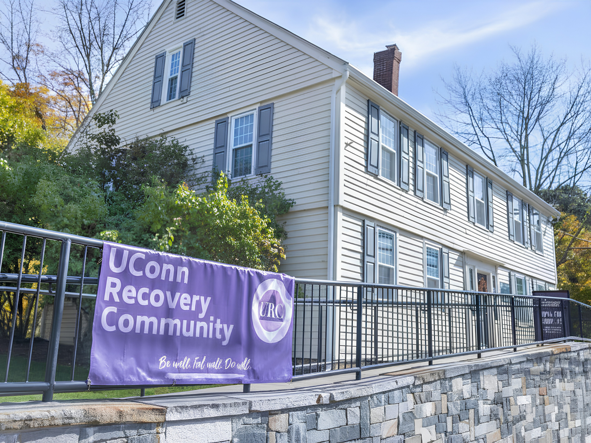 Purple “UConn Recovery Community” banner displayed on a railing in front of a light-colored house with trees and a clear sky in the background.