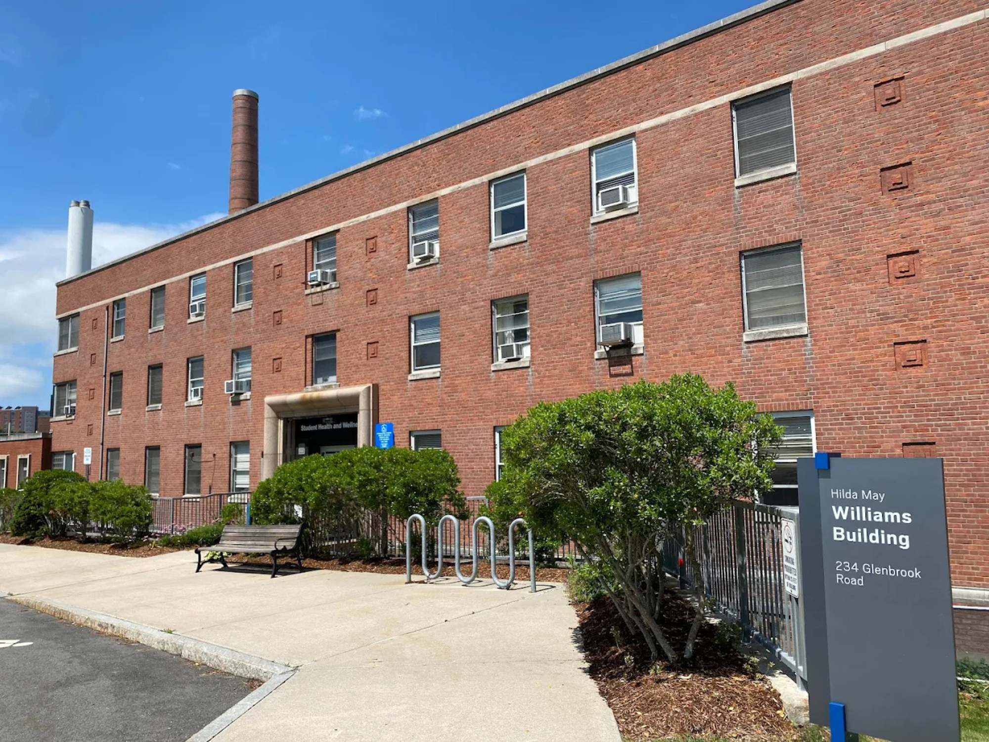 Exterior of Hilda May Williams Building with entrance, bench, bike rack, and surrounding greenery visible.