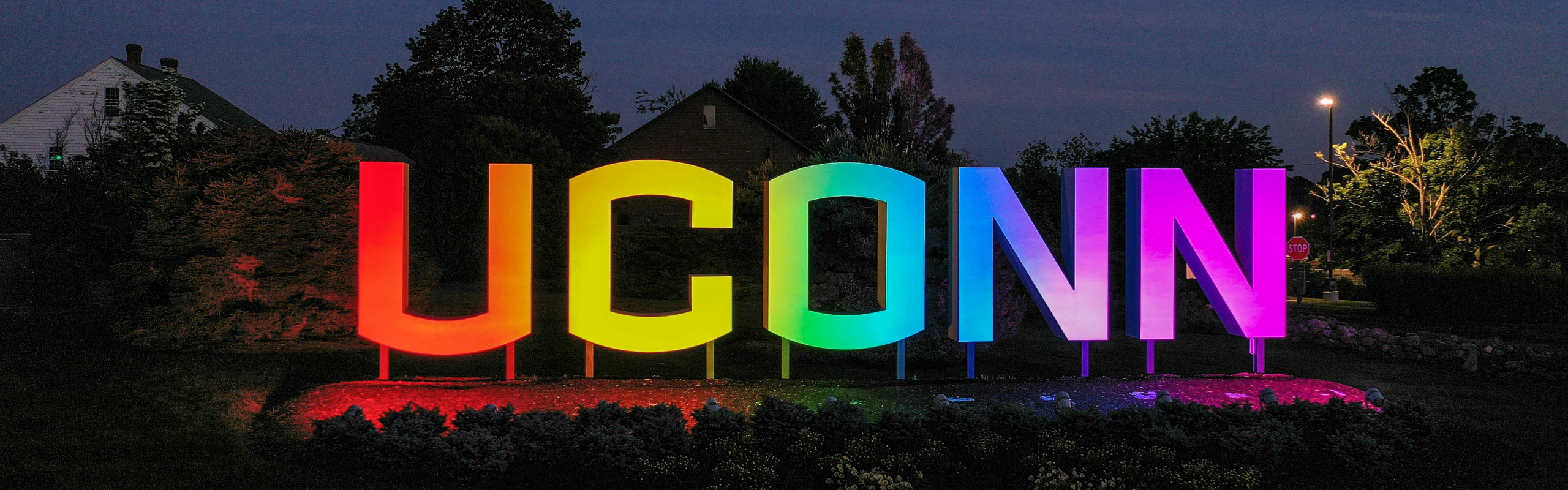 Large illuminated “UCONN” sign glowing in rainbow colors at night on a landscaped campus lawn.