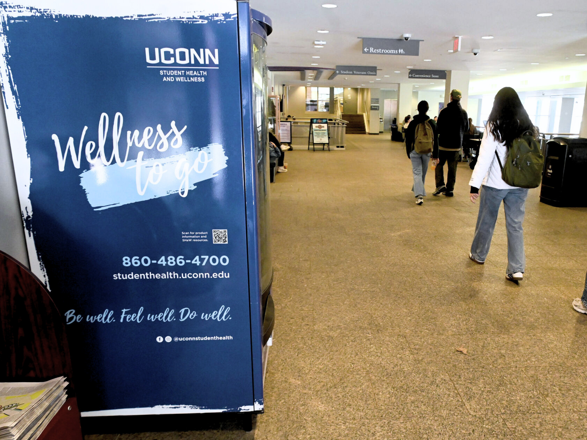 UConn Student Health and Wellness “Wellness to go” sign displayed in a campus hallway with students walking by.