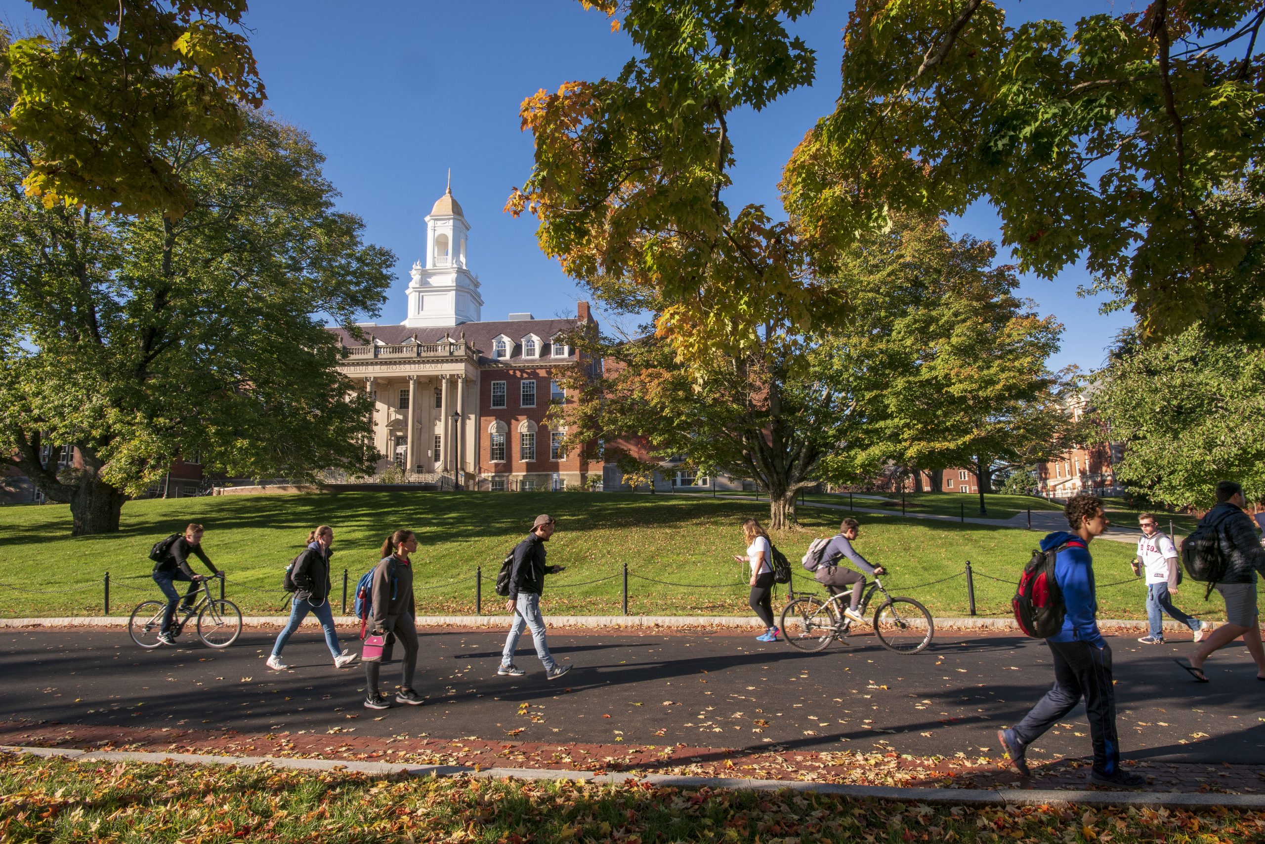 Students walk and bike along a campus path in front of a large academic building, surrounded by trees with fall foliage under a clear sky.
