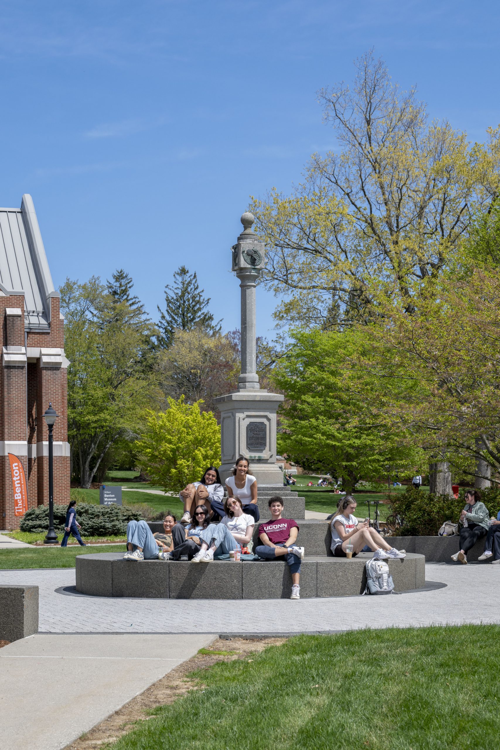 A diverse group of students relaxing and socializing on the curved stone steps of the "UConn Husky" statue plaza at the University of Connecticut Storrs campus on a sunny spring day.