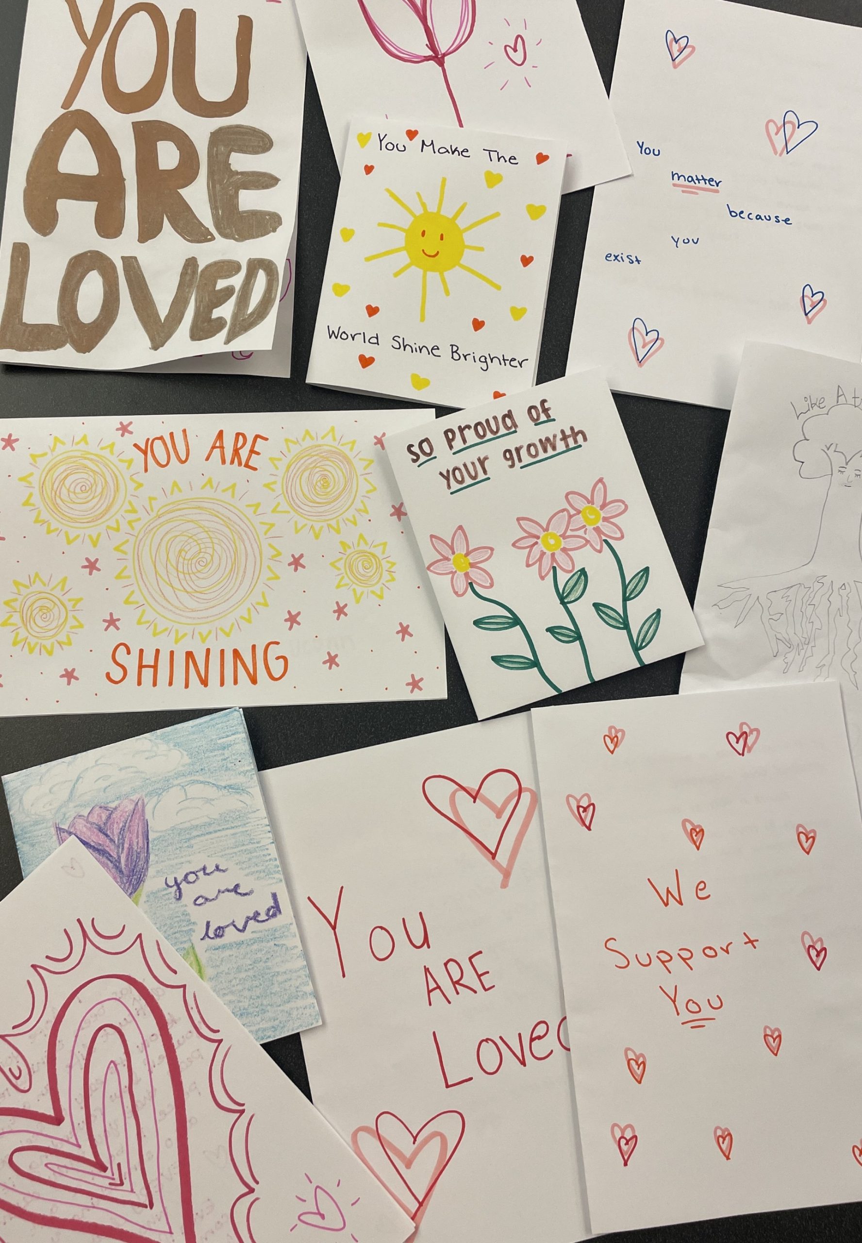 A flat lay collection of handmade cards and notes arranged on a dark tabletop. The cards feature colorful marker and colored pencil illustrations of suns, flowers, and hearts, accompanied by handwritten uplifting messages including "YOU ARE LOVED," "You Make The World Shine Brighter," "So proud of your growth," "YOU ARE SHINING," and "We Support You."