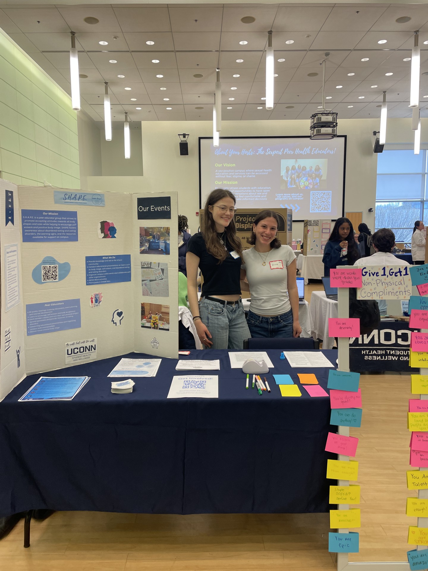 Two female students stand smiling behind an information table covered in a dark blue tablecloth at an indoor campus fair. To the left, a tri-fold poster board labeled "S.H.A.P.E." displays mission statements, event photos, and a UConn Student Health and Wellness logo. The table holds informational flyers, QR code sheets, and colored markers. On the right foreground post, a display board titled "Give 1, Get 1 Non-Physical Compliments" is covered in colorful sticky notes with encouraging handwritten messages like "You are deserving!" and "You are Epic." In the background, a large screen projects a slide titled "About Your Hosts: The Sexpert Peer Health Educators!